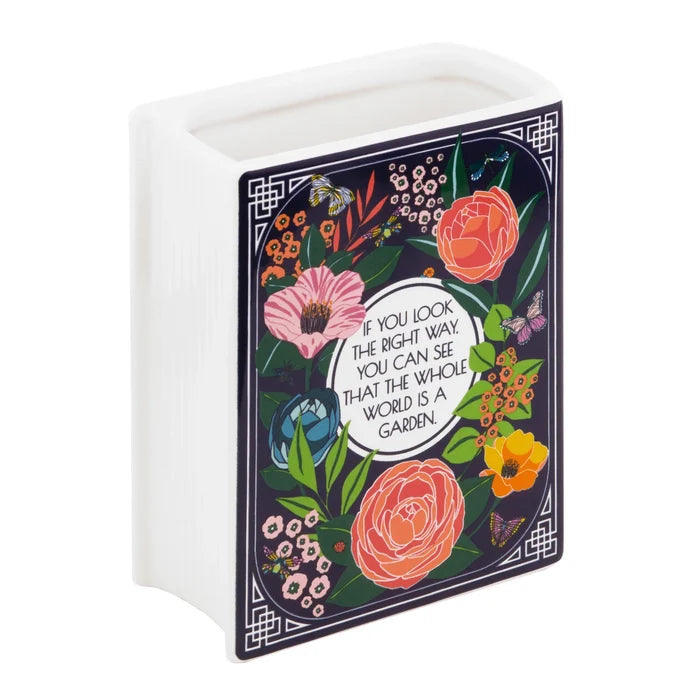 The back of a book-shaped white ceramic vase featuring a dark blue panel illustrated with colorful floral arrangements, butterflies, and a central quote that reads, "IF YOU LOOK THE RIGHT WAY, YOU CAN SEE THAT THE WHOLE WORLD IS A GARDEN."