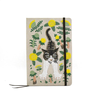 A hardcover notebook with a beige linen cover featuring an embroidered design of a black and white cat surrounded by yellow flowers and green leaves, secured with a black elastic band.