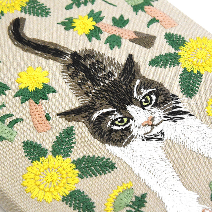 Close-up of a woven textile featuring a detailed embroidered-style cat face and body in black, white, and gray, surrounded by yellow flowers, green leaves, and peach carrots on a beige background.