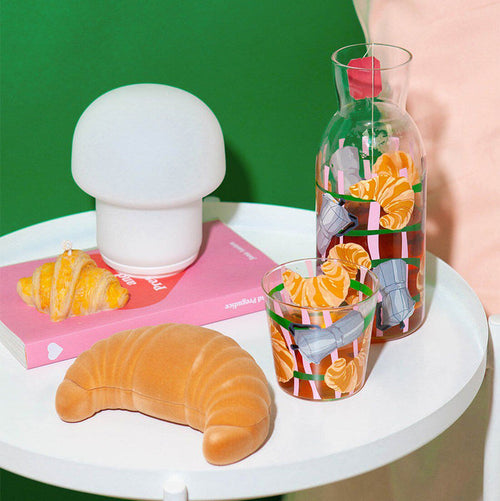 A clear glass carafe jar and a matching drinking glass on a small white table, featuring an illustrated pattern of moka pots and flaky croissants.