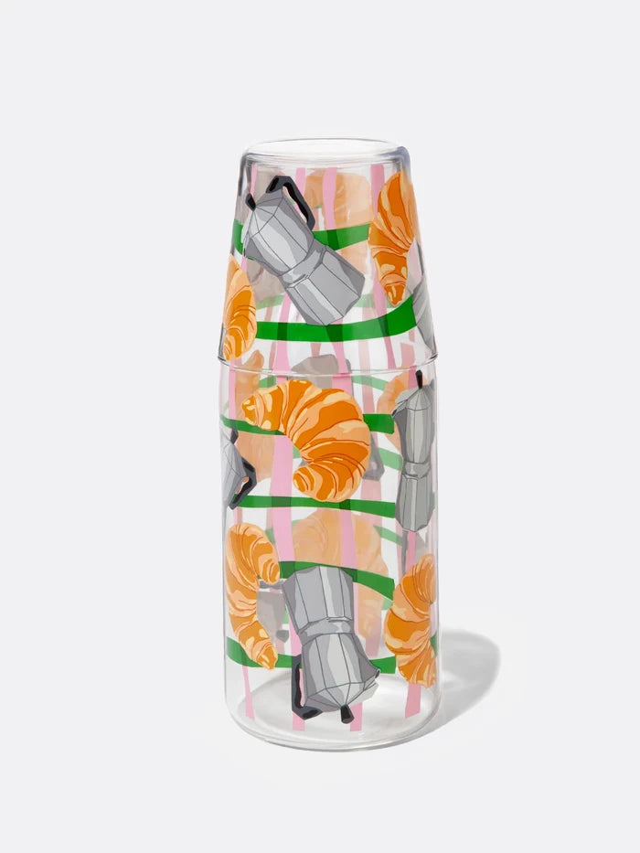 A clear glass carafe jar and a matching drinking glass, decorated with a graphic illustration of croissants and grey moka pots, isolated against a plain white background.