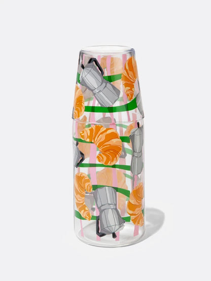 A clear glass carafe jar and a matching drinking glass, decorated with a graphic illustration of croissants and grey moka pots, isolated against a plain white background.