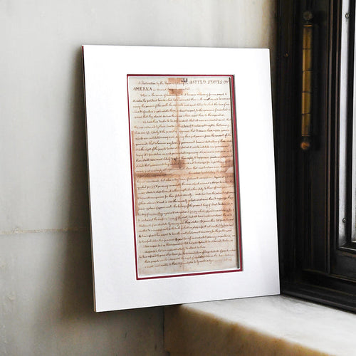 A matted replica print of the United States Declaration of Independence with an aged, parchment appearance and red trim, displayed upright inside a white photo matte against a marble sill.