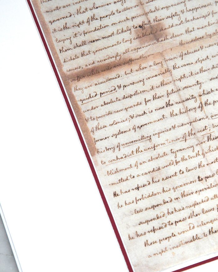 A close-up, angled view showing the handwritten cursive script on an aged, sepia-toned replica print of the United States Declaration of Independence, set within a red border.