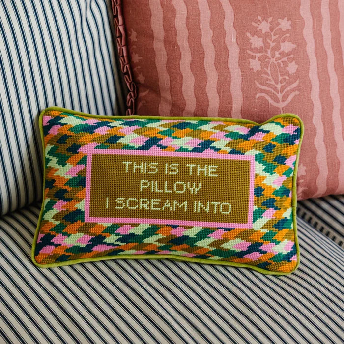 A rectangular needlepoint throw pillow featuring a multi-colored geometric zig-zag pattern and a central olive-green block with the text "THIS IS THE PILLOW I SCREAM INTO."