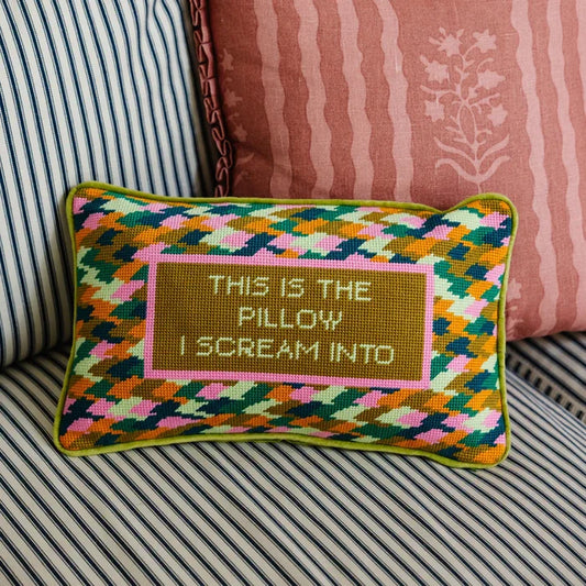 A rectangular needlepoint throw pillow featuring a multi-colored geometric zig-zag pattern and a central olive-green block with the text "THIS IS THE PILLOW I SCREAM INTO."